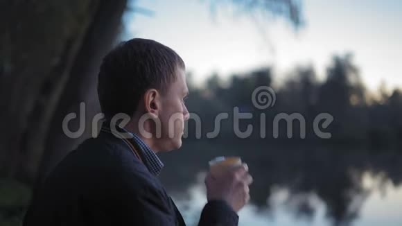 近景男喝热茶穿着紧身衣看太阳落山坐在森林的湖边视频的预览图