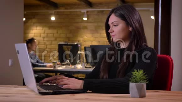一张正面的成年黑发女商人在笔记本电脑上打字的特写照片转向相机微笑着视频的预览图