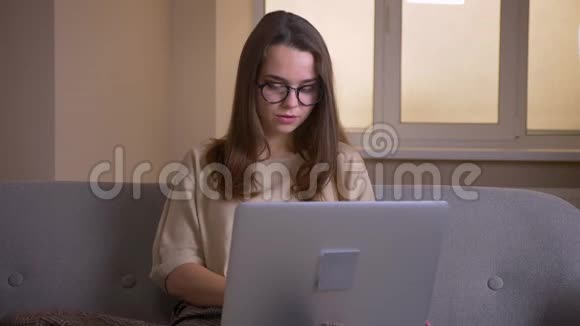 一位年轻漂亮的高加索女商人戴着眼镜坐在沙发上用笔记本电脑打字视频的预览图