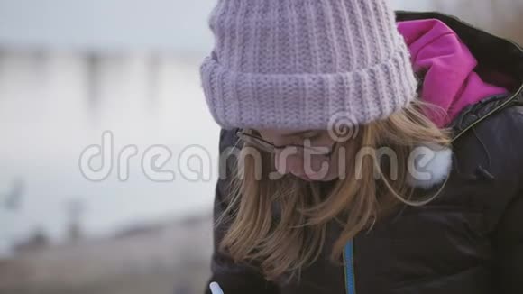 一个年轻的女孩戴着眼镜戴着一顶粉红色的帽子在一个春天的下午坐在河边用笔在笔记本上记笔记视频的预览图