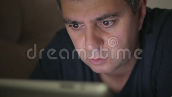 一个穿着蓝色T恤的男人躺在沙发上看着电子平板电脑脸上的恐惧和惊讶视频的预览图