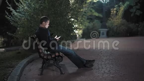严肃的男人在晚上坐在城市公园的长凳上用智能手机读信息视频的预览图