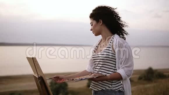 一位迷人的卷曲女艺术家在湖边的户外画架上画着她的画早晨柔软视频的预览图
