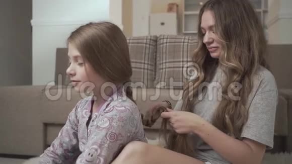 姐姐梳着小女孩的头发坐在家里沙发附近的地板上蓬松的地毯上姐妹关系视频的预览图