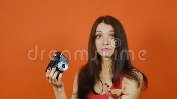 美丽的棕色头发的年轻女孩手里拿着相机情感一幅橙色背景的画室里的艺术肖像视频的预览图