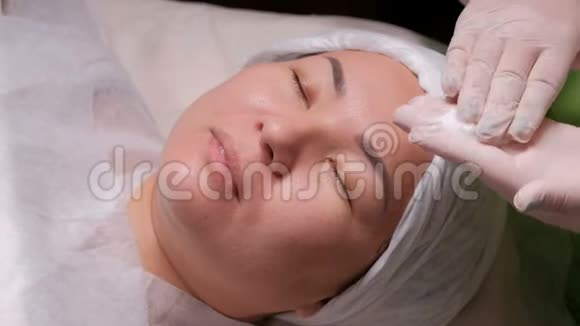戴着白手套的专业美容师在一个亚洲女人的脸上涂上一层滋养霜化妆程序视频的预览图