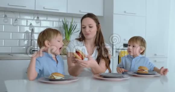 一位年轻漂亮的母亲穿着白色连衣裙带着两个孩子微笑着在厨房里吃着新鲜的汉堡快乐视频的预览图