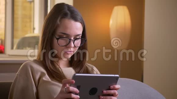 戴眼镜的年轻白种人女性的特写肖像使用平板电脑坐在舒适的室内沙发上视频的预览图