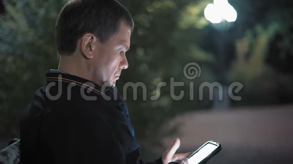 看新闻的严肃男人晚上坐在城市公园的智能手机上视频的预览图