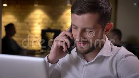 特写一位年轻开朗的白种人商人坐在笔记本电脑前微笑着笑着在电话中交谈视频的预览图