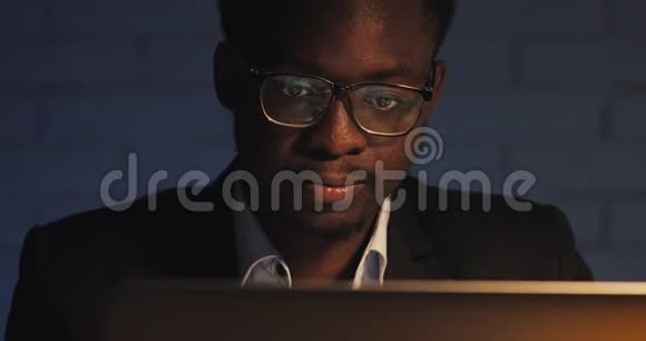 疲惫的年轻黑人商人在夜间办公室用笔记本电脑工作在黑暗房间加班的自由职业者视频的预览图