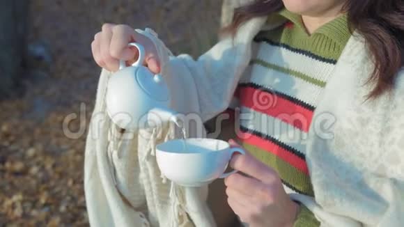 坐在长凳上的一位穿格子花格子的贴身美女用瓷茶壶倒绿茶用白杯子倒饮料视频的预览图