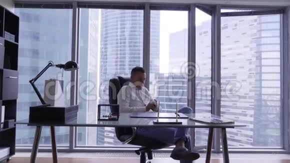 穿着白色衬衫的英俊黑发男子坐在现代办公室的玻璃办公桌后面视频的预览图
