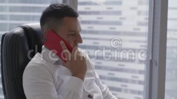 穿着白色衬衫的漂亮黑发男人在现代办公室的智能手机上讲话视频的预览图