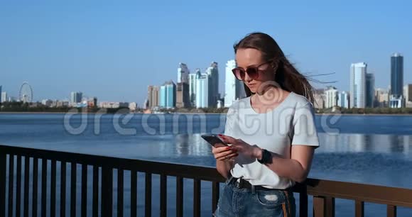 美丽的女孩长头发戴太阳镜使用智能手机应用程序在日落河码头附近4K视频的预览图