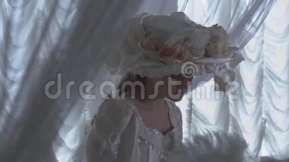 穿着白色老式球裙和戴着白色帽子站在透明窗帘的背景上的漂亮年轻女孩视频的预览图