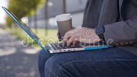 一个英俊的白种年轻人穿着夹克和围巾坐在长凳上手提电脑和咖啡在上面打字视频的预览图