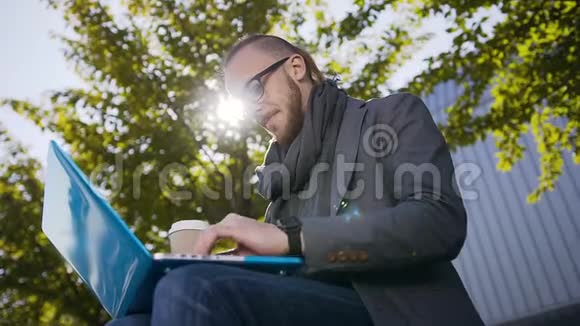 一张英俊的白人年轻人的肖像他留着胡子坐在长凳上拿着笔记本电脑戴眼镜走路的男孩视频的预览图