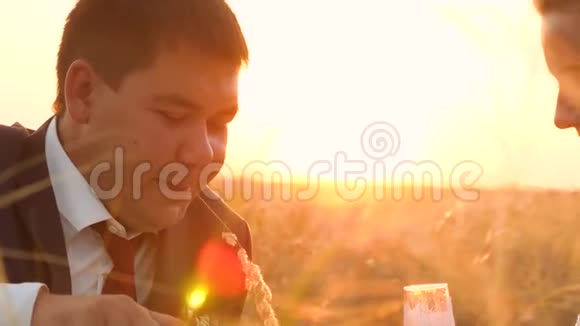 新娘和新郎被倒入香槟杯中坐在草地小穗在日落恋爱中的浪漫夜晚视频的预览图