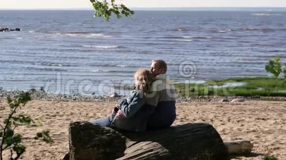 一位孕妇正在湖边拥抱她的丈夫坐在原木上年轻家庭视频的预览图