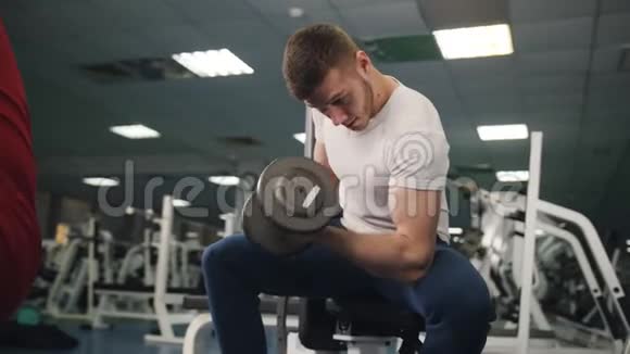 在健身房里男人坐着用一只手举起哑铃年轻的健美运动员借助视频的预览图