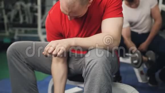 在健身房里男人坐着用一只手举起哑铃年轻的健美运动员借助视频的预览图