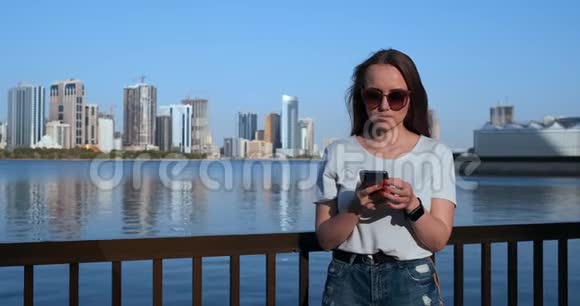 美丽的女孩长头发戴太阳镜使用智能手机应用程序在日落河码头附近4K视频的预览图