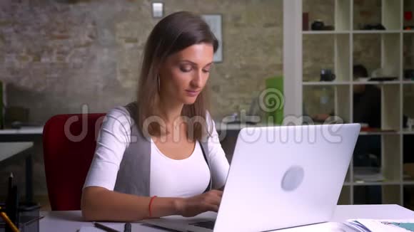 成年女性办公室工作人员正在笔记本电脑上打字并转向一台相机微笑着在室内工作场所视频的预览图