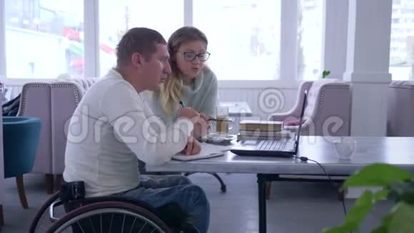 残疾发展女教师戴眼镜为轮椅上的残疾男性使用笔记本电脑进行讲座视频的预览图