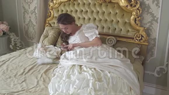 年轻女子穿着球服躺在金色装饰的床上用手机发短信女孩用小工具视频的预览图