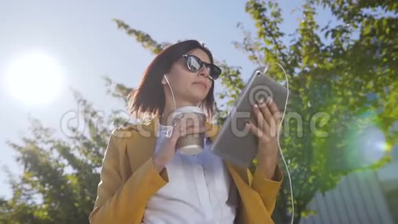 美丽的年轻女子戴着太阳镜拿着热咖啡在杯子里同时她在平板电脑上发送信息户外活动视频的预览图