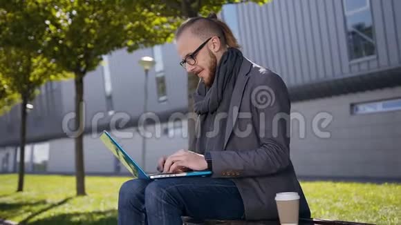 一张英俊的白人年轻人的肖像他留着胡子坐在长凳上拿着笔记本电脑帅哥男学生视频的预览图