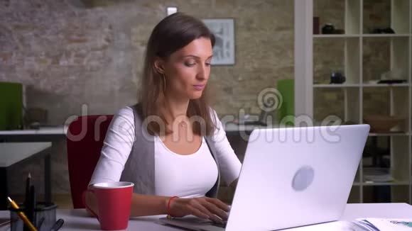 漂亮的女办公室职员在笔记本电脑上打字喝着咖啡她的工作变得很有活力笑容满面视频的预览图