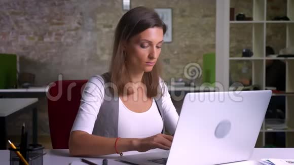成年女性办公室工作人员在笔记本电脑上打字把视线转向室内工作场所的摄像机视频的预览图