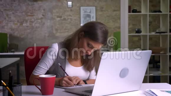 漂亮的办公室工作人员坐在笔记本电脑前在室内工作场所做笔记喝咖啡视频的预览图