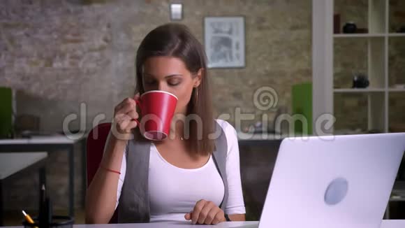 漂亮的女办公室职员坐在笔记本电脑前喝着美味的咖啡对着相机微笑视频的预览图