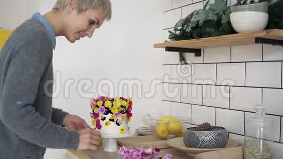 在现代的白色厨房里留着短发的女人用五颜六色的花朵装饰着小白蛋糕视频的预览图