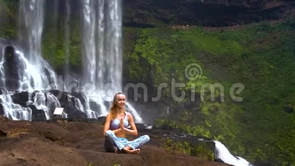 金发女孩坐在瀑布的岩石上的普拉纳亚瑜伽姿势视频的预览图