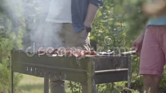 在花园里做烧烤的不可辨认的男人的特写镜头夏日休闲朋友们在烤架上煮肉视频的预览图