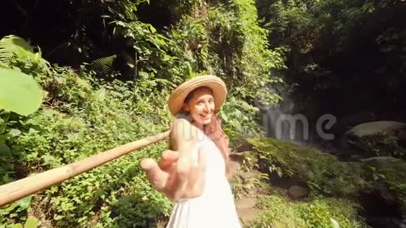 年轻的微笑混合种族女孩穿着白色连衣裙和草帽牵着手带领人们走向神奇的丛林瀑布生活方式视频的预览图