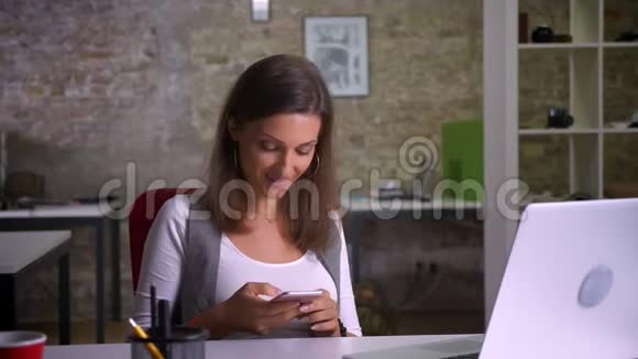 漂亮的女办公室工作人员坐在笔记本电脑前在电话上发短信笑了起来视频的预览图