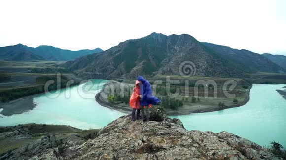 两个穿着滑稽雨衣的恋人站在山的边缘手牵手背面视频的预览图