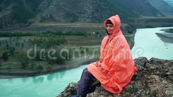 穿着橙色雨衣的女孩坐在卡屯河边的山顶上美丽的风景视频的预览图