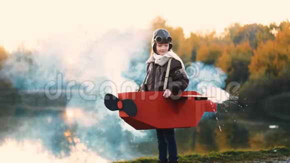 快乐的小飞行员女孩沿着日落湖在有趣的纸板飞机与彩烟玩飞行员慢动作视频的预览图