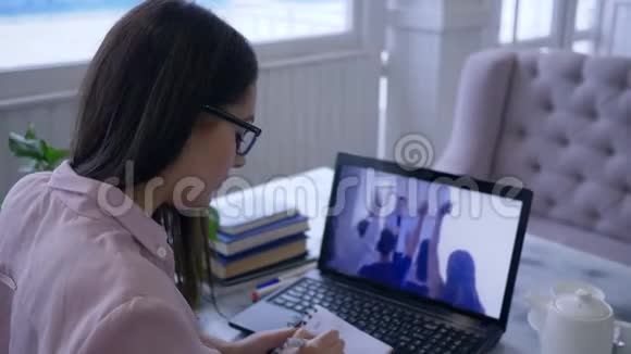 互动培训女学生戴眼镜在笔记本上看视频时用笔写在记事本上视频的预览图