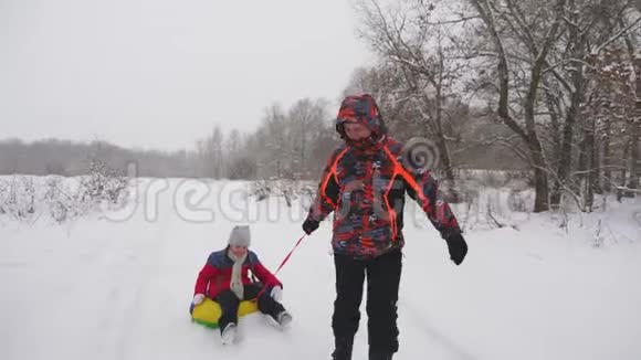 快乐的爸爸骑着一个孩子在白色的雪道上雪橇圣诞节假期成人和儿童的有趣游戏这就是视频的预览图