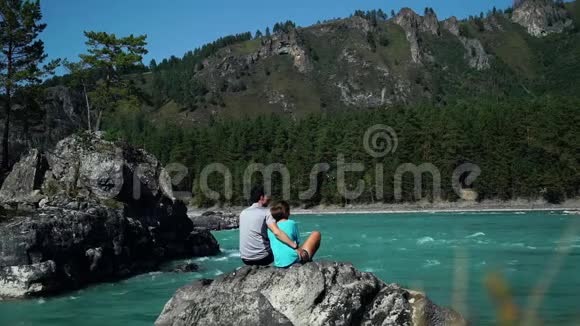 年轻的一对夫妇坐在快山河边男朋友拥抱一个女孩背面视频的预览图