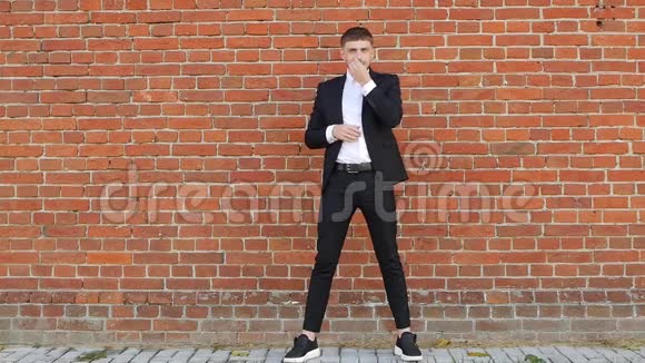 一个穿着黑色西装的白种人年轻人站在砖墙上抚摸着他的胡子和胡须慢着抄着视频的预览图