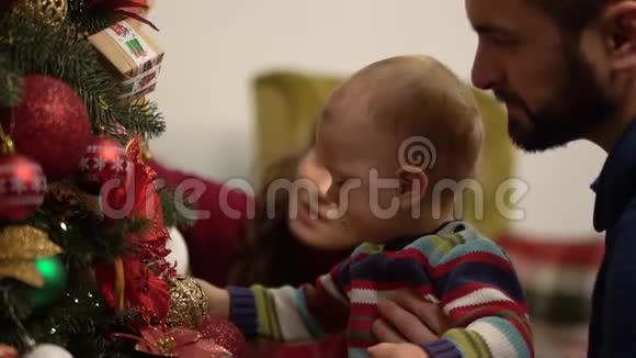 母亲父亲和小宝宝坐在装饰圣诞树的近照处男子抱着孩子靠近杉树展示视频的预览图