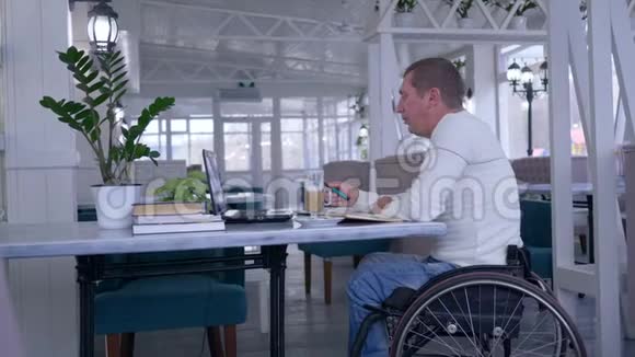 在线课程中坐在轮椅上的残疾学生使用笔记本电脑端着书坐在桌子旁喝咖啡视频的预览图